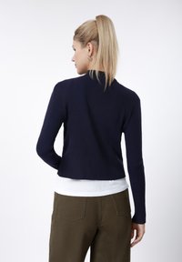 Marineblå ribbestrikket genser med lange ermer og høy hals, kort modell. Brukt over en hvit skjorte og kombinert med olivengrønne bukser.