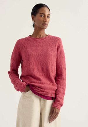 Donna che indossa un maglione girocollo rosa testurizzato sopra una camicia rosa scuro, abbinato a pantaloni beige chiaro, in piedi contro uno sfondo chiaro.