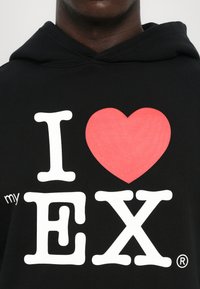 Fekete kapucnis pulóver nagy fehér felirattal: "I", egy piros szív, valamint vastag, fehér "EX" betűk. A felirat bal oldalán egy kis "my" is látható.