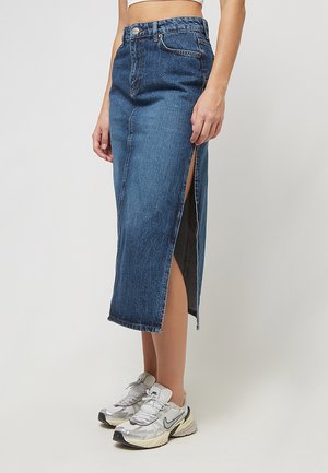 Frau trägt einen hoch taillierten blauen Denim-Midirock mit seitlichem Schlitz, kombiniert mit weißen und grauen Sportsneakern.