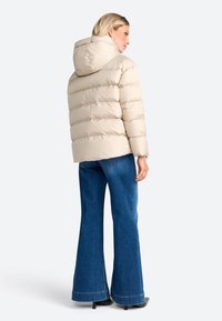 Beige pufferjack met capuchon, met doorstikte delen en een gladde textuur, gecombineerd met uitlopende blauwe jeans en zwarte hakken.