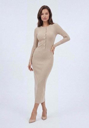 Kobieta stojąca w dopasowanej beżowej sukience midi z prążkowanego materiału, z długimi rękawami, guzikami z przodu oraz beżowymi, spiczastymi szpilkami na jednolitym jasnym tle.
