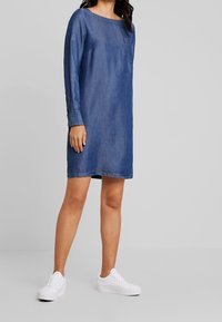 Tummansininen farkkuasu, jossa on pitkät hihat, pyöreä pääntie ja rintatasku. Yhdistetty valkoisiin lenkkareihin.