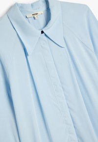 Camisa azul claro de botones hecha de tela suave. Presenta un gran cuello, tapeta delantera y un botón negro en contraste.