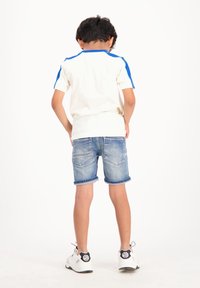 Vit skjorta med blå detaljer, denimshorts med uppvikta benslut och vita sneakers med svarta detaljer. Sedd bakifrån.