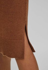 Maglione marrone a maglia con una texture morbida, caratterizzato da uno spacco laterale e dettagli a coste sul fondo. Il tessuto appare caldo e accogliente.