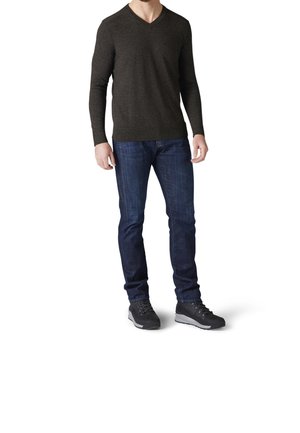 Mann trägt dunkelgrauen V-Ausschnitt Pullover, blaue Jeans und schwarze Wanderschuhe, steht vor weißem Hintergrund, Kopf nicht sichtbar.