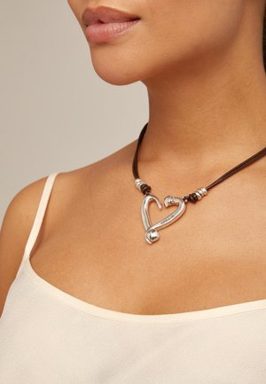 Colgante en forma de corazón de plata en un cordón de cuero marrón con acentos de plata, llevado sobre piel desnuda. Superficie lisa y reflectante con un diseño elegante.