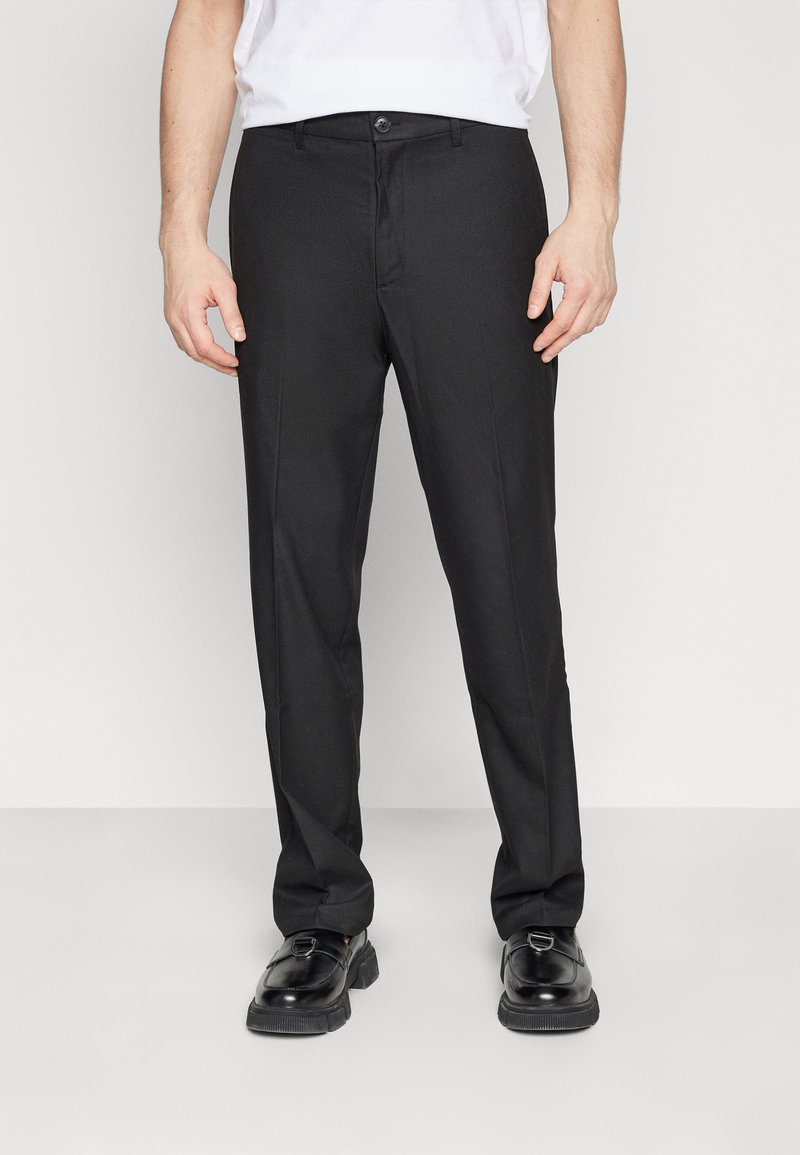 Zwarte pantalon met een rechte snit, gladde textuur, platte voorkant en een enkele knoopsluiting. Draagt een wit T-shirt en zwarte schoenen.
