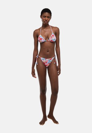 Femme debout pieds nus portant un bikini deux pièces à imprimé floral avec un col halter et un bas noué sur les côtés, sur un fond uni.