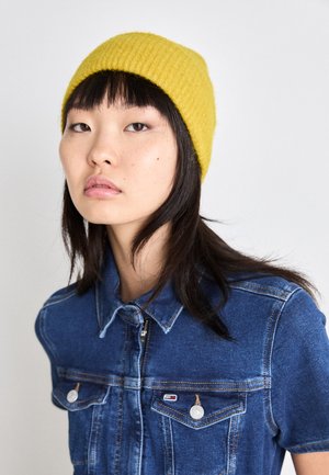 Joven con el pelo negro y liso que lleva un gorro de punto amarillo y una chaqueta de mezclilla azul, frente a un fondo blanco liso.