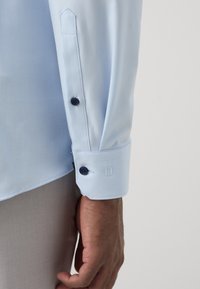 Hellblauer Hemdärmel mit schwarzen Knöpfen, getragen von einer Person mit brauner Haut, kombiniert mit hellbeiger Hose vor einfarbigem Hintergrund.