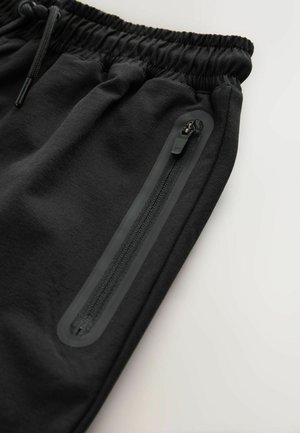 Črne trenirka hlače z elastičnim pasom, z zadrgo ob strani, izdelane iz gladkega blaga z matirano površino.