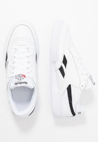 Weiße Sneaker mit schwarzen Akzenten und strukturiertem Leder, ausgestattet mit perforierten Details, flachen Schnürsenkeln und einem Logo auf der Zunge.