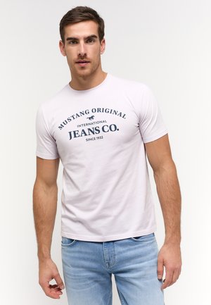 Egy férfi világos lila Mustang Original Jeans Co. pólót és világoskék farmert visel, egy sima fehér háttér előtt állva.