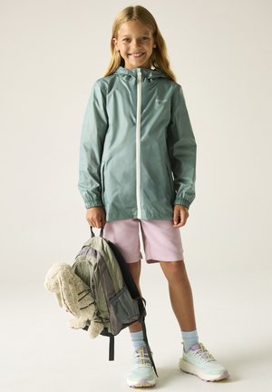 Smiling girl wearing a green rain jacket and pink shorts holds a gray and green backpack with a stuffed bunny toy hanging from it.