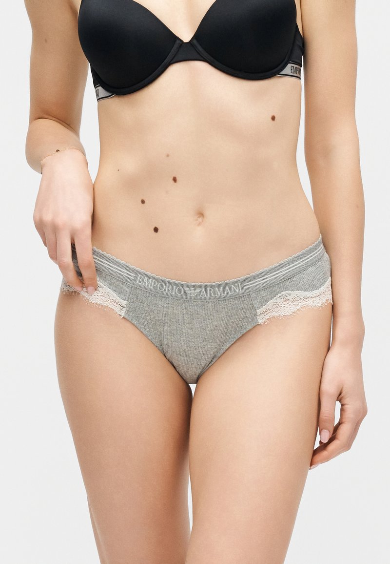 Γυναίκα που φοράει γκρι εσώρουχο Emporio Armani με δαντέλα και μαύρο σουτιέν, με ορατές ελιές στο σώμα της, σε ουδέτερο φόντο.