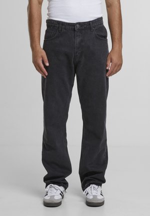 Homme portant un jean noir, un débardeur blanc et des baskets blanches avec des rayures noires, debout devant un fond gris uni.