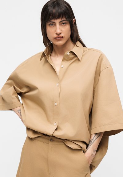 Νεαρό άτομο με μεσαίου μήκους σκούρα μαλλιά φοράει υπερμεγέθη μπεζ πουκάμισο και ταιριαστό παντελόνι, στέκεται με το ένα χέρι στην τσέπη, με ουδέτερη έκφραση.