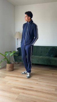 Marineblå træningssæt med hvide striber, lynlåslukning jakke og bukser med snor. Bæres med grønne og hvide sneakers. Indendørs setting med en grøn sofa.