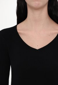 Femme portant un haut noir à col en V, montrant le cou, les clavicules et le haut de la poitrine avec trois petits grains de beauté sur la peau.