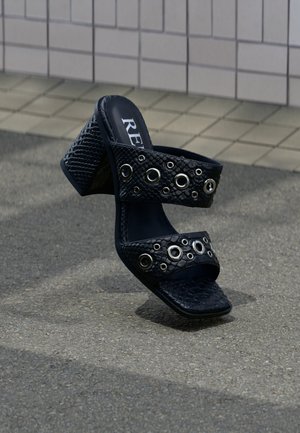 Μαύρο σανδάλι με τακούνι και υφασμάτινα λουριά με ασημένια τρουκς, τοποθετημένο σε εξωτερικό δάπεδο κοντά σε πλακιδωτό τοίχο.