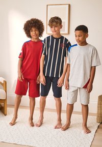 Trois garçons debout pieds nus en tenue décontractée : un t-shirt rouge avec un short assorti, une chemise marine rayée avec un short foncé, et un t-shirt gris avec un short clair.