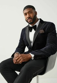 Marineblauwe fluwelen blazer met satijnen revers, wit overhemd, zwarte vlinderdas en een gestreepte pochet; zittend op een zwarte stoel.