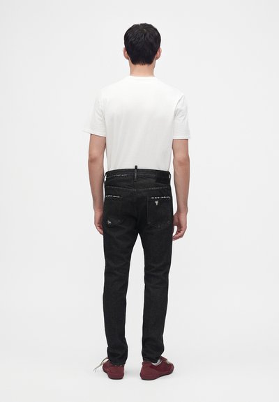 Homme portant un t-shirt blanc uni, un jean noir déchiré et des baskets rouges, debout de dos sur un fond blanc uni.