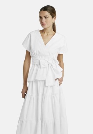 Frau mit braunen Haaren in einem weißen V-Ausschnitt-Kleid mit kurzen Ärmeln, einer großen Taillenschleife und einem gestuften, knöchellangen Rock, die seitlich blickt.