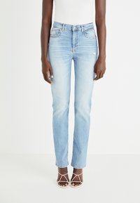 Jeans de denim azul claro con corte recto, tiro alto, cinco bolsillos y detalles de desgaste sutil. Combinados con sandalias blancas de tiras.