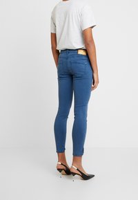 Person, der eng anliegende blaue Jeans und schwarze Slingback-Absätze trägt, steht vor einem schlichten hellen Hintergrund und blickt von der Kamera weg.
