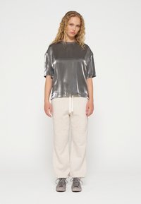 Metallisch silbernes T-Shirt mit kurzen Ärmeln, kombiniert mit hellbeigen Sweatpants mit Kordelzug. Strukturierter Stoff, lockere Passform und lässiger Stil.