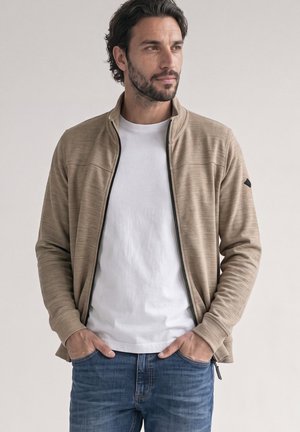 Mann mit dunklen Haaren und Bart, der eine beige Reißverschlussjacke über einem weißen T-Shirt und blauen Jeans trägt, mit den Händen in den Taschen, schaut zur Seite.