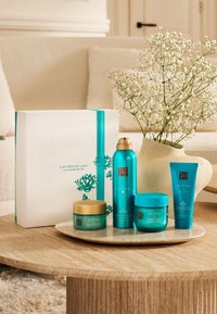 Zestaw czterech turkusowych produktów do pielęgnacji skóry, wyeksponowanych na okrągłym drewnianym stole, z złotymi akcentami i białym pudełkiem prezentowym z turkusową wstążką.