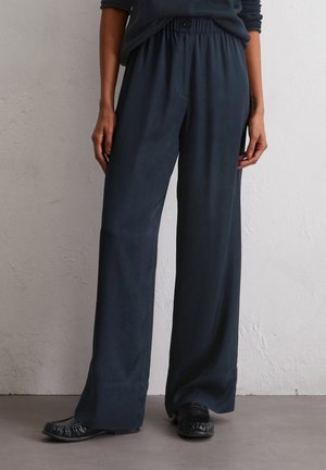 Dunkelblaue, weit geschnittene Hosen mit glatter Textur, elastischem Bund und einem Knopfdetail. Kombiniert mit schwarzen, flachen, gerafften Schuhen.