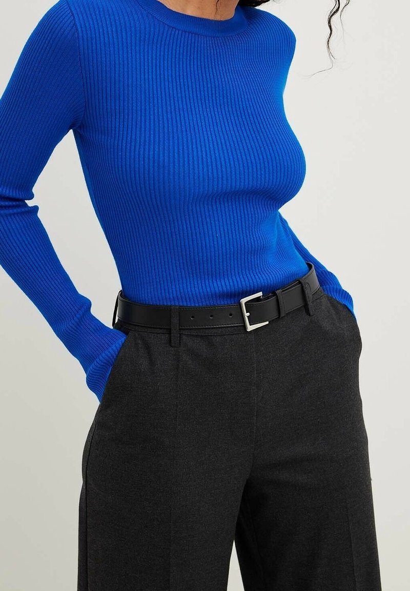 Kobieta w jasnoniebieskim, prążkowanym topie z długim rękawem, włożonym do wysokich, szarych spodni z czarnym paskiem. Ręce w kieszeniach.