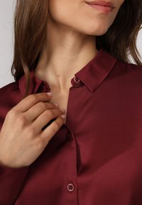 Chemise en satin bourgogne avec un col à boutons, deux boutons assortis et une texture lisse. Main effleurant délicatement la zone du col.