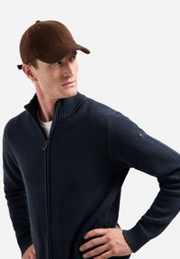 Marineblauwe gebreide zip-up trui met een textuurpatroon en hoge kraag, gecombineerd met een bruine vilten baseballpet met een verstelbare band aan de achterkant.
