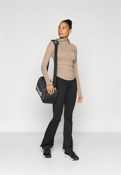 Μπεζ μακρυμάνικη μπλούζα με γιακά μισής φερμουάρ, συνδυασμένη με μαύρο παντελόνι. Το άτομο κρατάει μια μαύρη τσάντα με λευκό λογότυπο. Μαύρα αθλητικά παπούτσια.