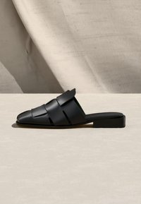 Μαύρο δερμάτινο μοκασίνι χωρίς κορδόνια με πλεκτό σχέδιο λουριού και χαμηλό τακούνι σε απλή μπεζ επιφάνεια.