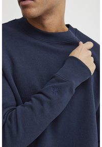 Marine crewneck sweatshirt i myk bomullsmiks, med ribbestrikkede mansjetter og et lite brodert emblem på ermet. Glatt tekstur, avslappet design.