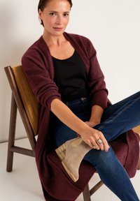 Vinröd stickad cardigan, svart topp, blå jeans och beige ankelstövlar med träklack. Sitter på en trästol med texturerad ryggstöd.