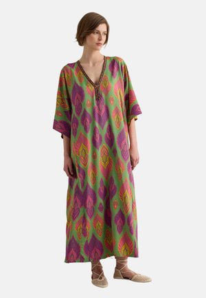 Woman wearing a long, loose green dress with large pink and yellow leaf patterns and beige lace-up flat shoes, standing against a white background.