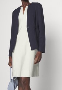 Veste marine ajustée avec un col rond et une fermeture à un bouton, portée sur une robe gris clair longueur genou, tenant un petit sac à main bleu.