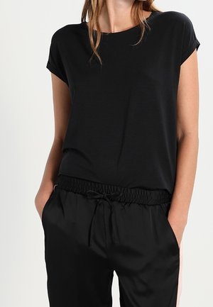 Vrouw in een zwart t-shirt met korte mouwen en zwarte elastische broek met koord, met handen in de zakken, tegen een eenvoudige achtergrond.