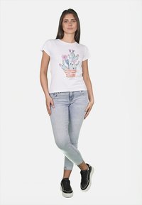 T-shirt bianca con una grafica di cactus e fiori, abbinata a jeans skinny blu chiaro e sneakers nere, in piedi su uno sfondo semplice.