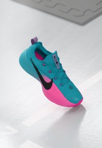 Turkis og pink træningssko med sort Nike swoosh, pink øjer og trækstrop, præsenteret på en reflekterende hvid overflade.