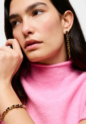 Kobieta w różowym swetrze z golfem, złota kwiatowa bransoletka na nadgarstku i pasujące długie złote kolczyki z motywem kwiatowym, zamyślona patrzy na bok.