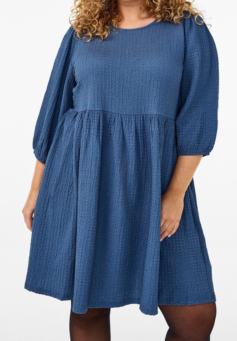 Personne aux cheveux bouclés portant une robe bleue texturée longueur genoux avec des manches ballon trois-quarts sur fond blanc.